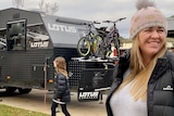A woman in a beanie smiles broadly as she stands in front of a caravan with bikes on the back.