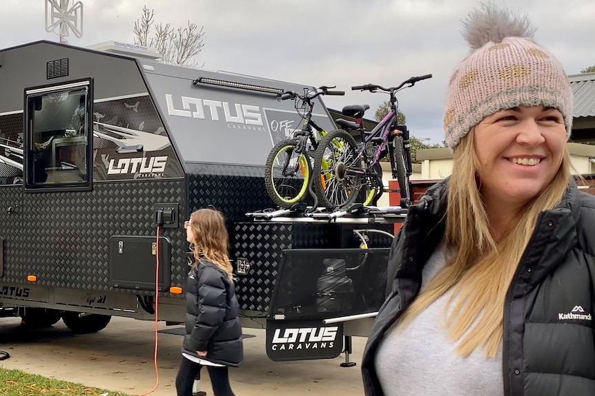 A woman in a beanie smiles broadly as she stands in front of a caravan with bikes on the back.