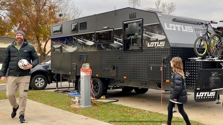 Un homme tient un ballon de football et une fille marche vers lui devant une caravane noire. 