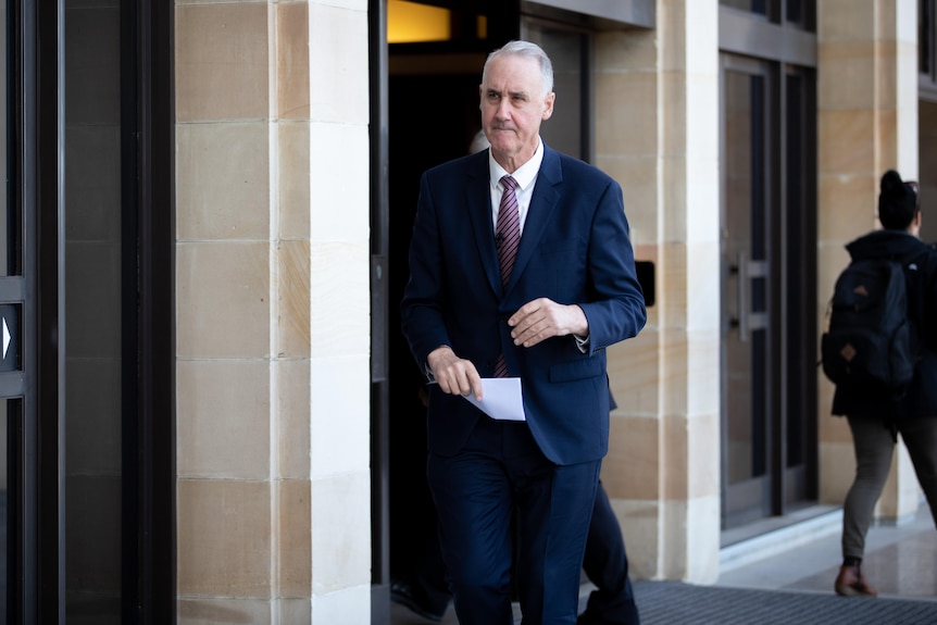 A man in a suit walks along next to a building holding a piece of paper.