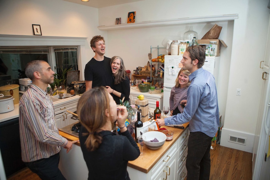 A group of young people stand around laughing and talking in a kitchen.