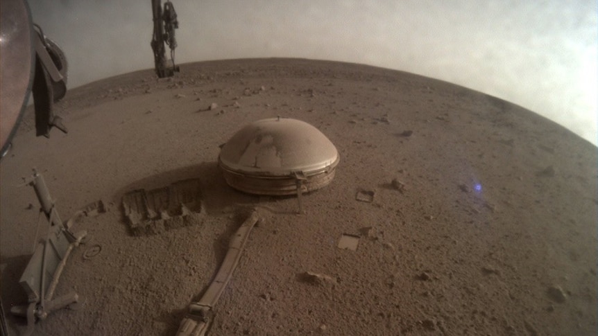 A fish-eye lens photo shows a small, dust-covered white dome sitting on the rocky, dirty surface of a strange planet.