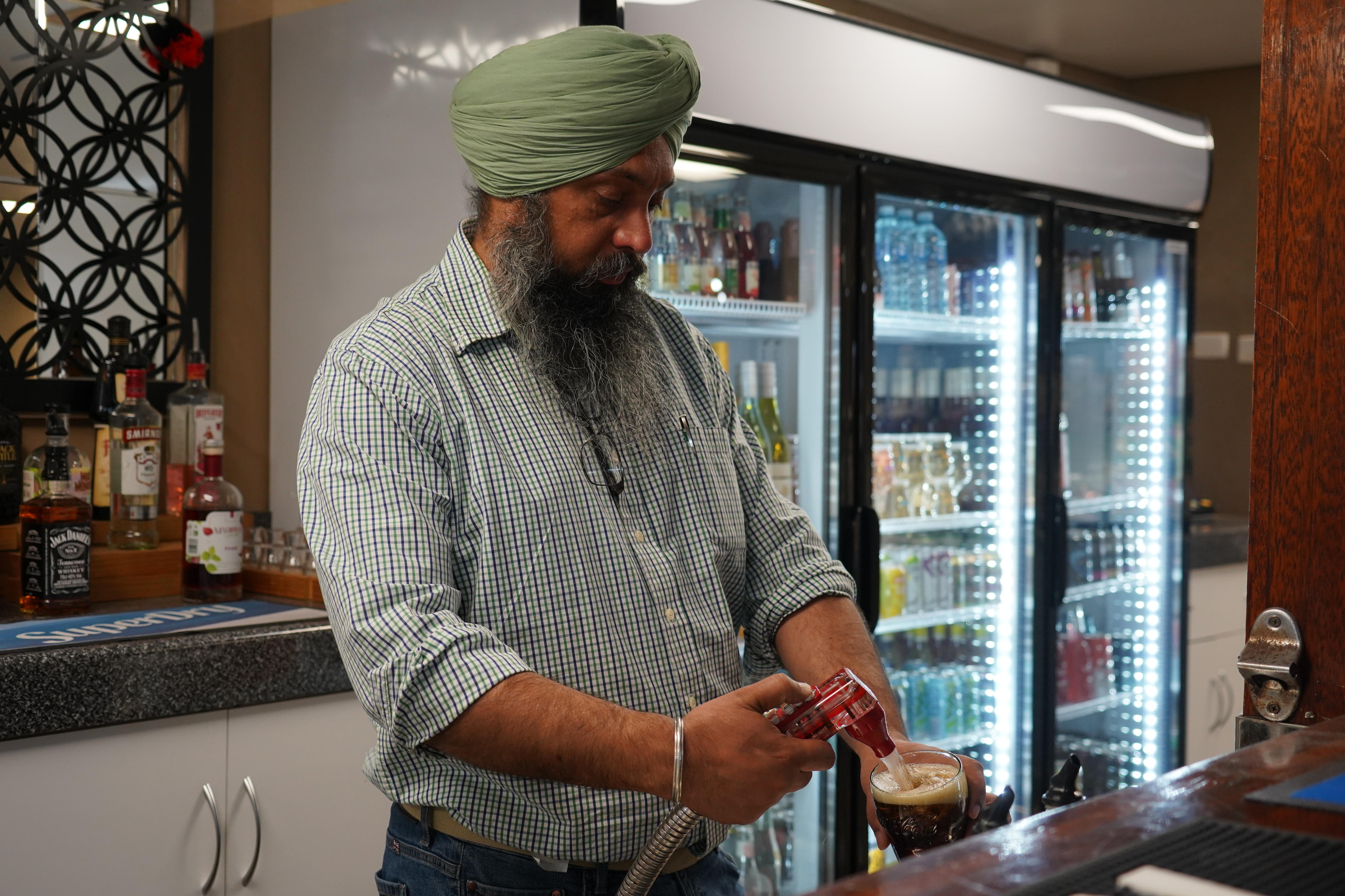 A man in a checked shirt and turban stands behind a bar pouring a drink. A fridge of drinks is in the background.