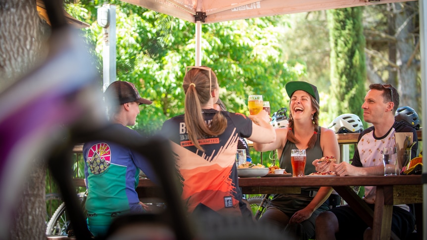 Four people sit at an outdoor table and cheers their drinks, surrounded by bikes and helmets. 