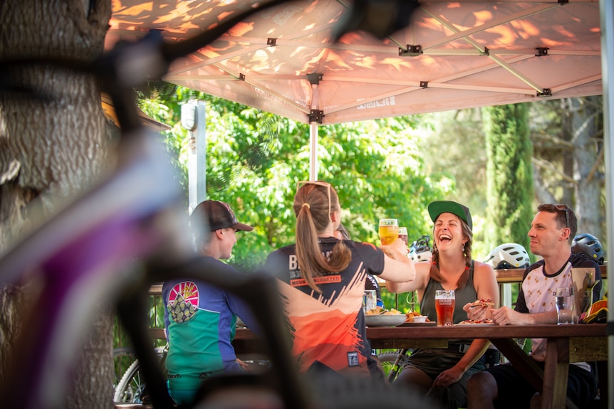 Four people sit at an outdoor table and cheers their drinks, surrounded by bikes and helmets. 