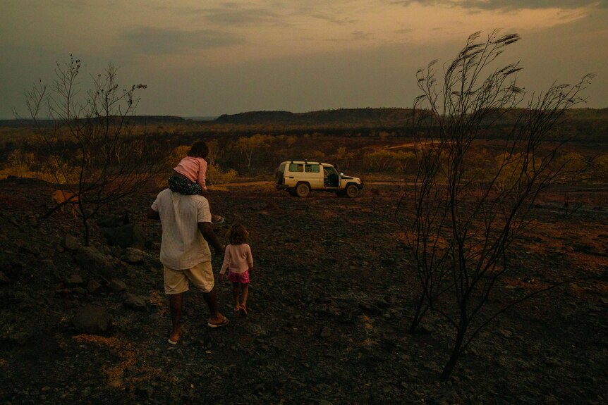 A man carries a girl on his shoulders as he walks towards a car 