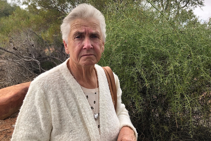 A woman with white hair wearing a white cardigan looks at the camera, frowning