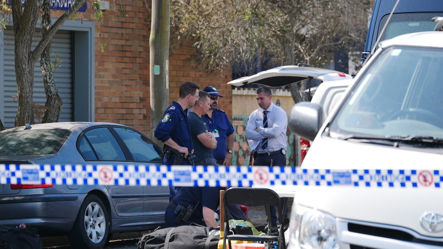 Four police officers, two in uniform and two in plain clothes, behind police tape at a crime scene.