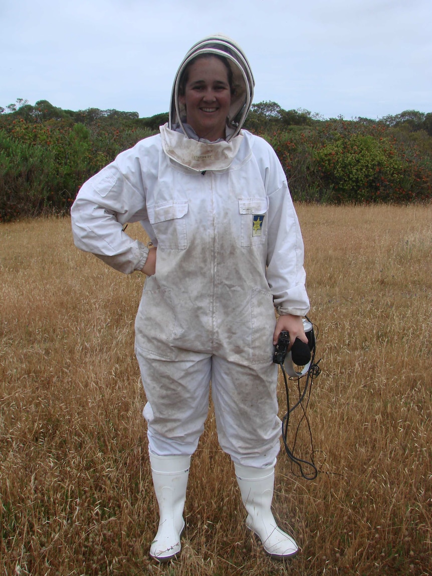 Woman in white overalls, boots and mesh head covering holding microphone and recording equipment while standing in a field.