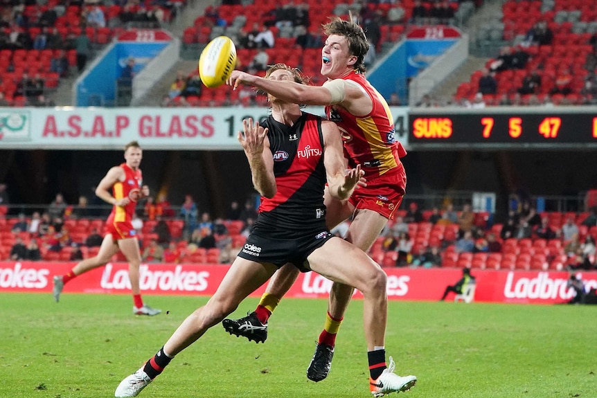 A Gold Coast Suns AFL player stretches out his left arm to punch the ball in front of an Essendon opponent.