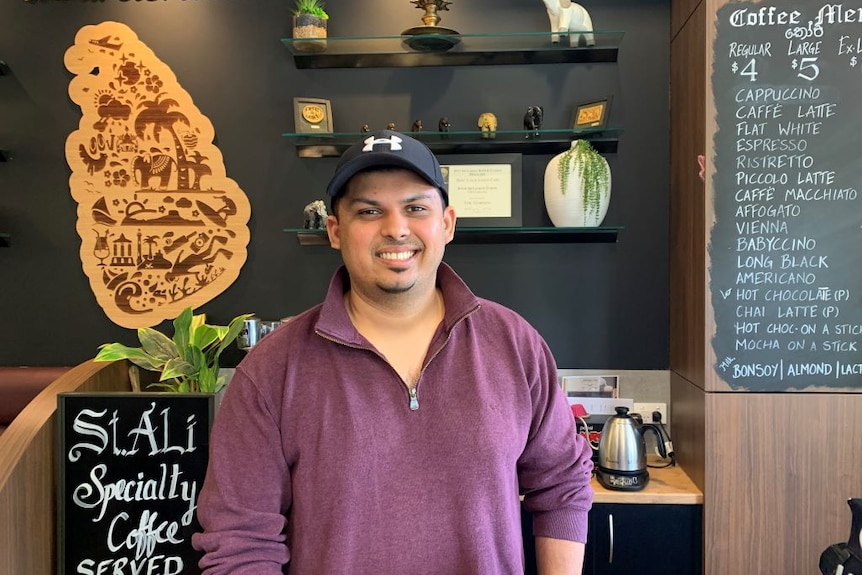 A man wearing a cap and red jumper stands in a cafe with a coffee menu and Sri Lankan trinkets on the wall behind him.