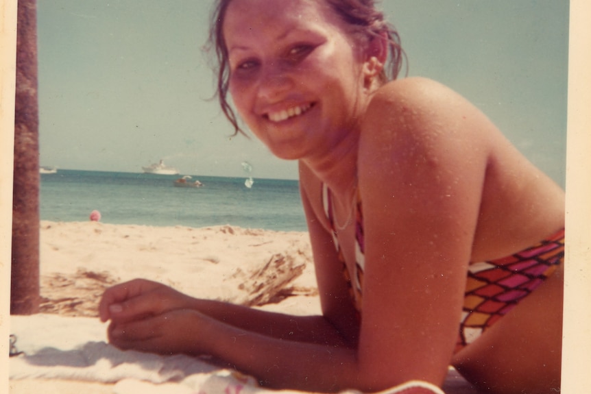 A young Indigenous woman in a bikini sunbakes on a beach.  She is smiling.  