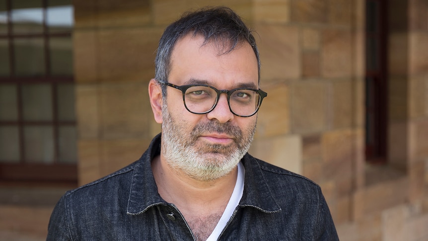 Colour photo of artist Asad Raza standing in front of sandstone bricked wall and windows.