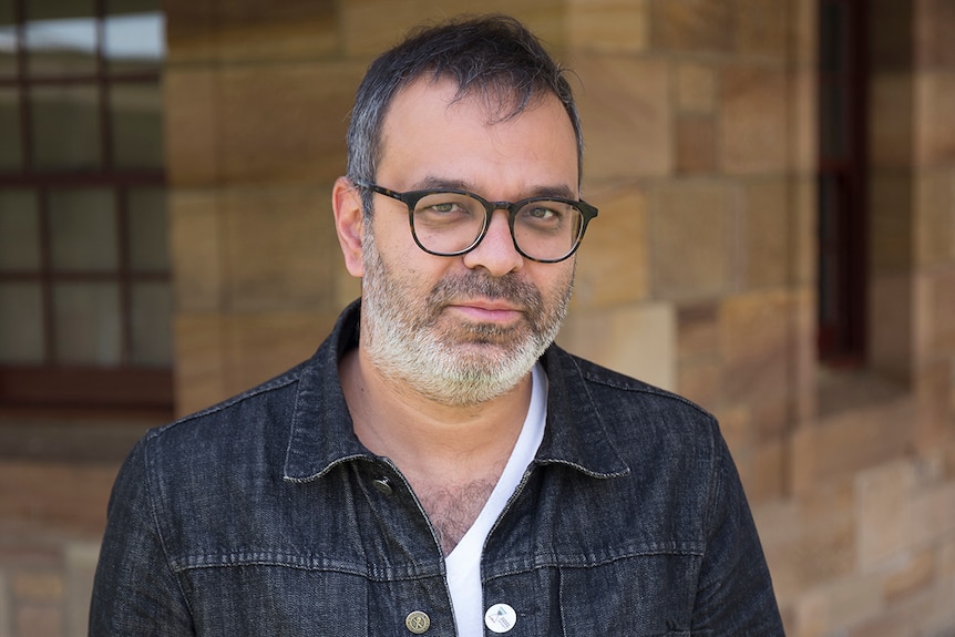 Colour photo of artist Asad Raza standing in front of sandstone bricked wall and windows.