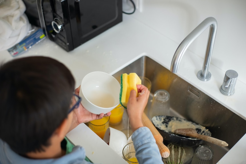 A boy rubs a bowl with a yellow and green scourer with a sink full of dirty dishes in front of him