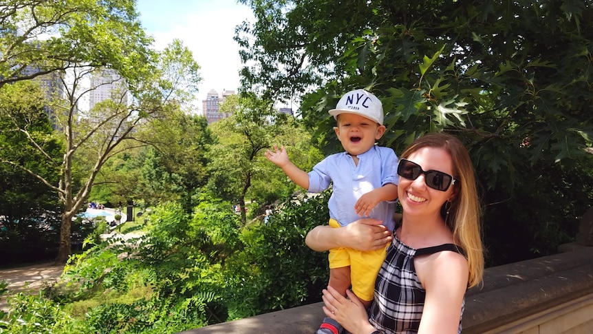 A blonde woman holds up a happy waving toddler wearing an NYC hat in front of trees and a city view.