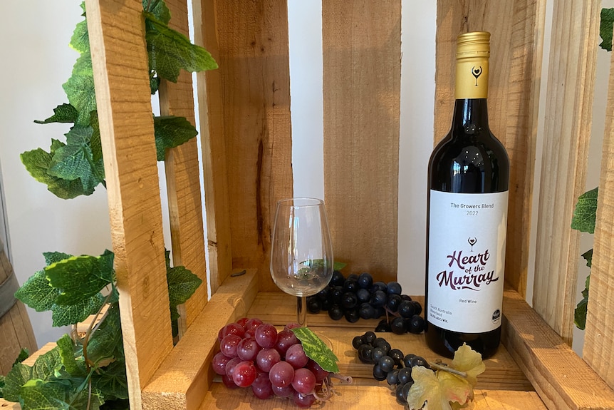 A bottle of Heart of the Murray wine in a wood box next to fake grapes and a wineglass.