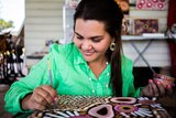 An young indigenous woman in a green linen shirt uses the back end of a paintbrush to create her art.