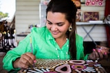 An young indigenous woman in a green linen shirt uses the back end of a paintbrush to create her art.