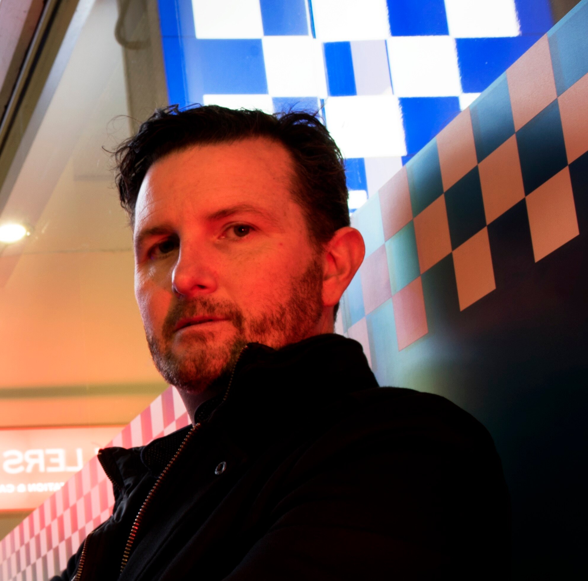 A man with red light on his face and ble and white chequers in the background. 