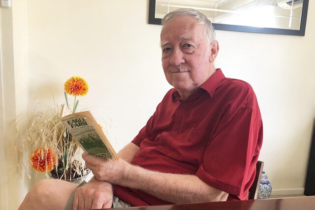 An older man wearing a red shirt and sitting down looking at a racing booklet with Eagle Farm printed in green on the cover.