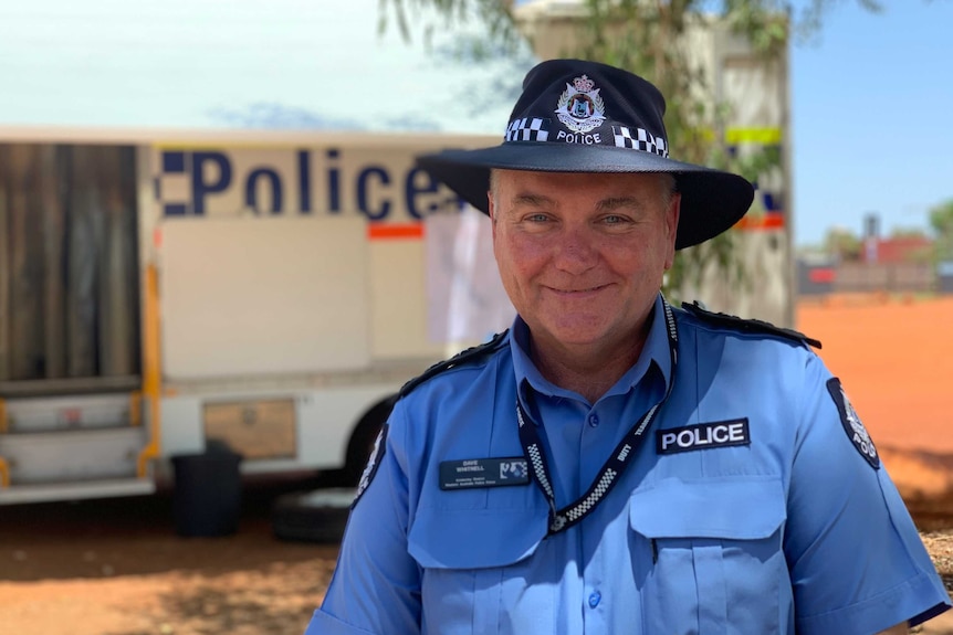 A smiling man in a police uniform stands in front of a WA police van and a blue sky