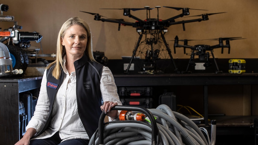 A woman sits on the edge of a large vacuum cleaner type object, drones can be seen in the background.