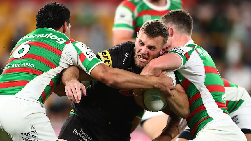 A Penrith Panthers NRL player holds the ball with his left hand as he is tackled by two South Sydney opponents.