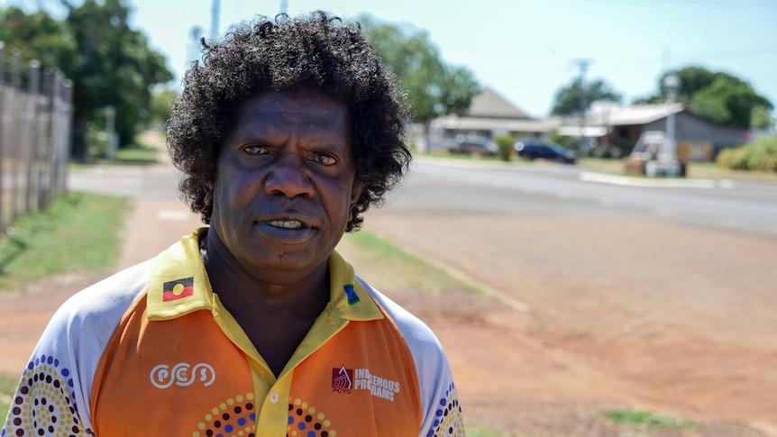 An Indigenous man stands outside in a rural area, smiling. 