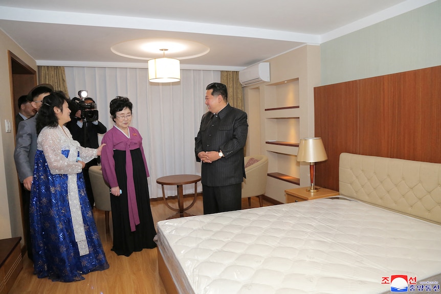 uśmiechnięty Kim Jong Un stoi ze splecionymi rękami przed grupą ludzi, którzy weszli do nowej sypialni