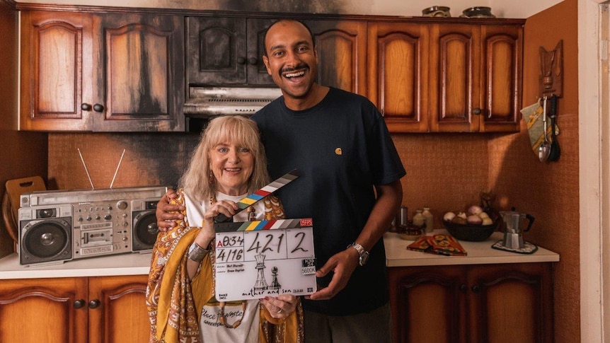 Mother and Son Denise Scott as Maggie Boye and Matt Okine as Arthur Boye standing in a kitchen. Scott is holding a clipboard.