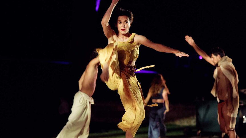 Four dancers on stage, black backdrop. In foreground, woman in sand-coloured clothes has one leg and two arms raised mid-dance.