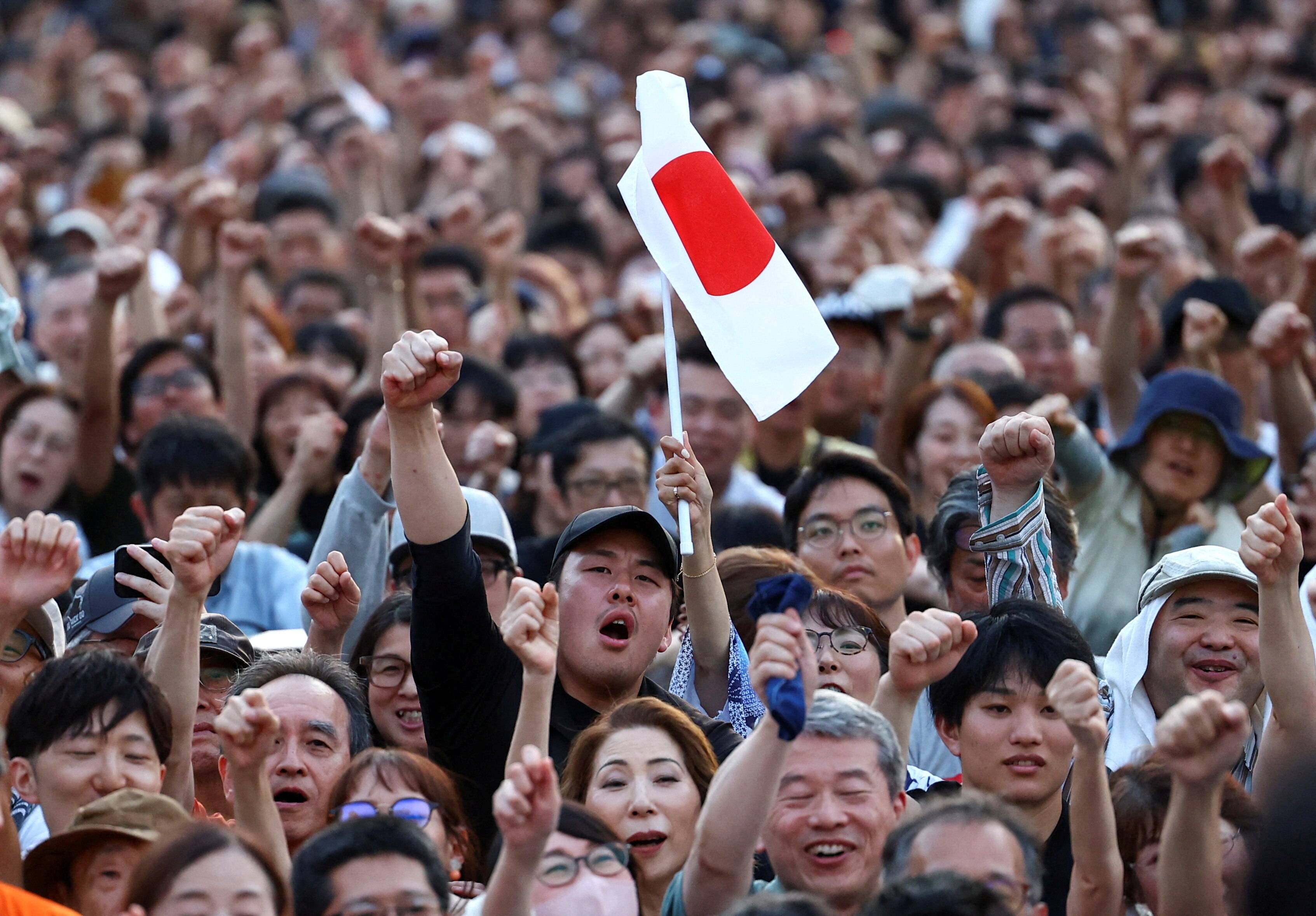 A crowd of people, one holding a Japanese flag and another cheering and pumping his fist.
