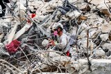 A rescuer wearing safety and breathing equipment moves through the waist-high rubble of a collapsed apartment building.
