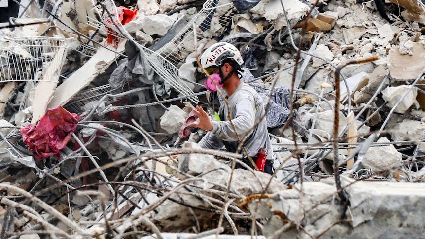 A rescuer wearing safety and breathing equipment moves through the waist-high rubble of a collapsed apartment building.