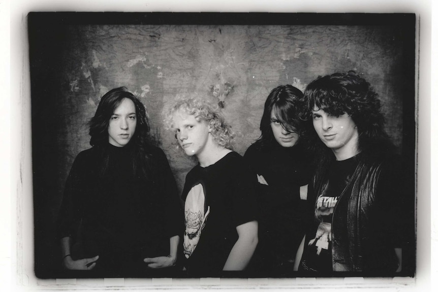 Four men wearing black t-shirts and jackets stand in front of a grey wall.