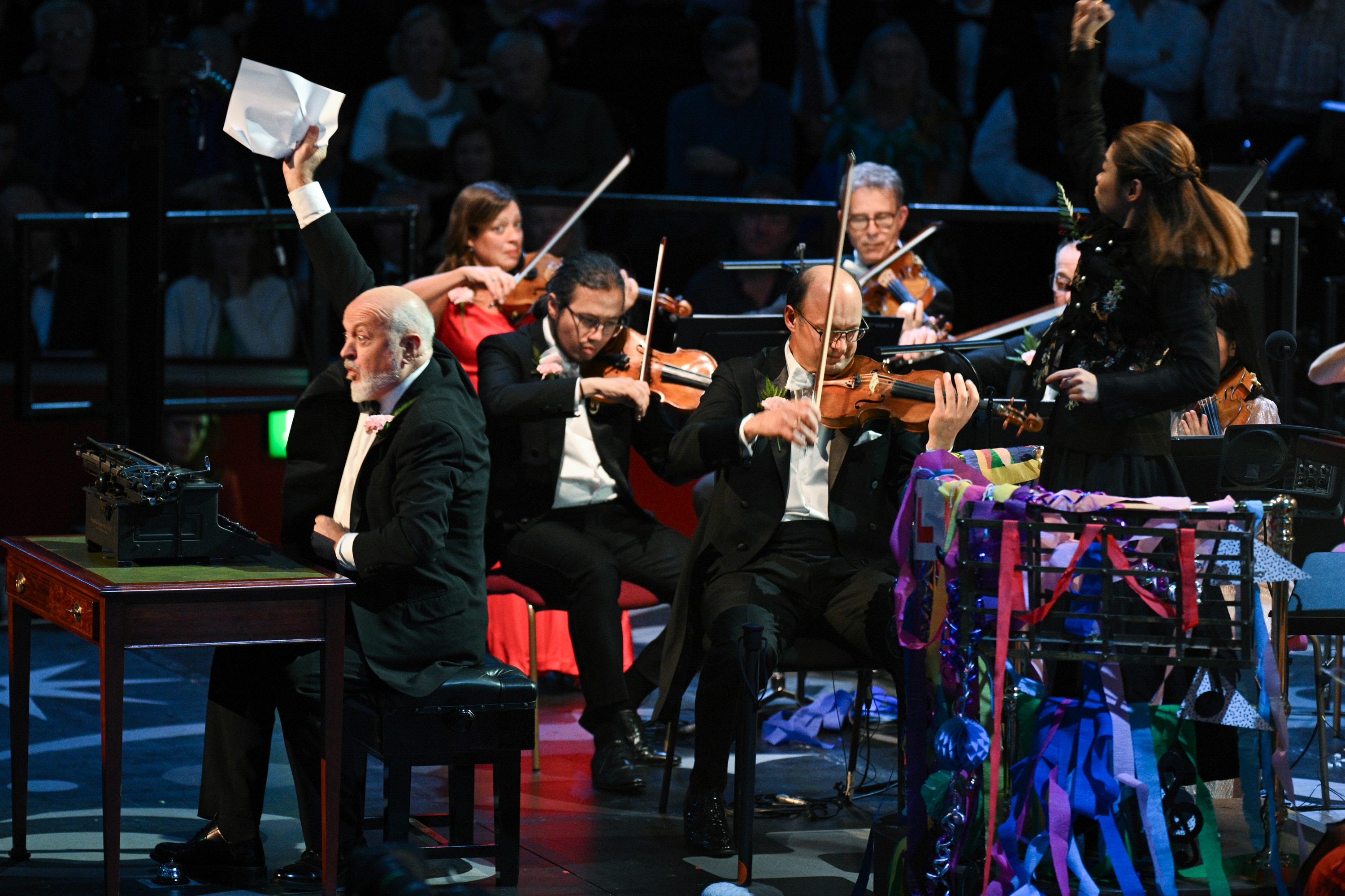 Bill Bailey raises a crumpled paper beside a typewriter as orchestra members play during the 2025 Last Night of the Proms.