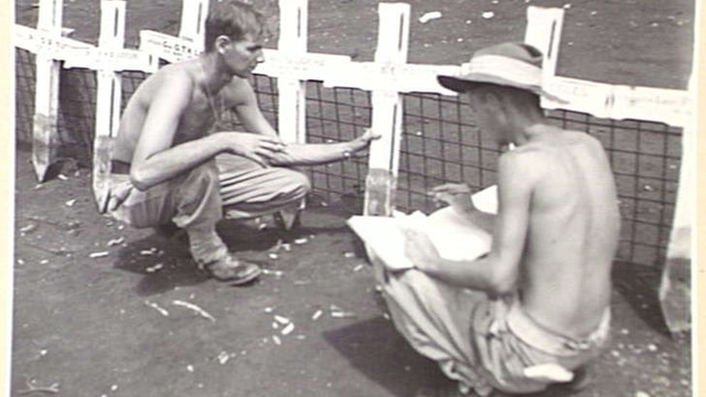 Two shirtless men crouching near wooden crosses, one blonde, other wearing a uniform hat, reading from a document.