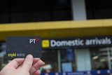 A hand holds a myki card, sign in background reads 'Domestic Arrivals'
