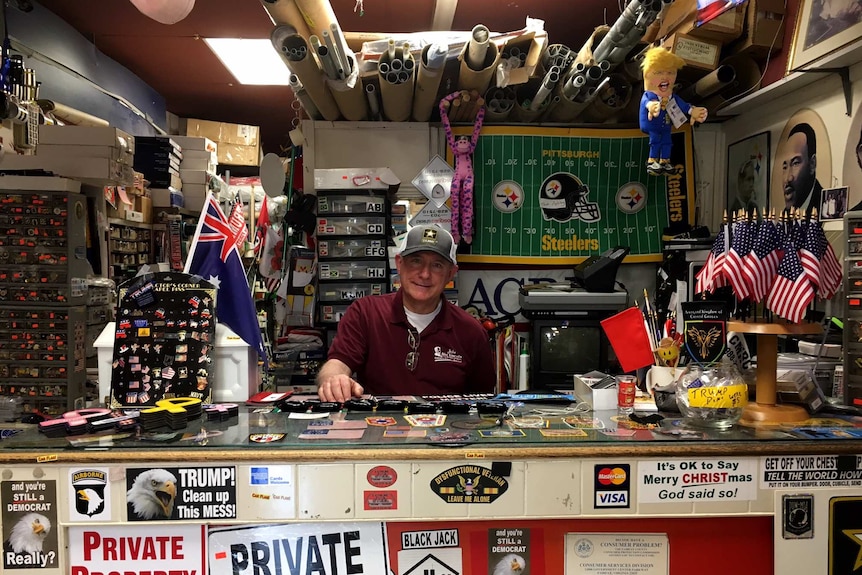 John Lipnicky sits behind a desk at a busy shopfront. There are Trump stickers and American flags all over his desk.