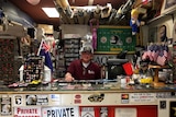 John Lipnicky sits behind a desk at a busy shopfront. There are Trump stickers and American flags all over his desk.