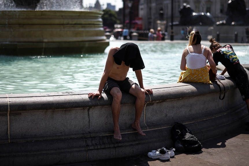 A man sits with his bare feet dangling off the side of a fountain. His head is covered from the sun with a T-shirt. 