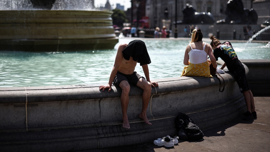 A man sits with his bare feet dangling off the side of a fountain. His head is covered from the sun with a T-shirt. 