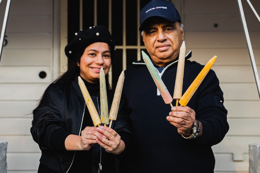 Shikha and Harbir smile on a front porch, holding long, rocket shaped ice cream sticks (kulfi) of different flavours.