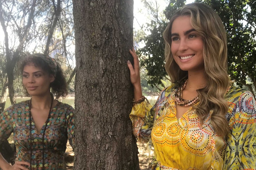 Two models stand among trees wearing silk dresses featuring Aboriginal dot paint prints.