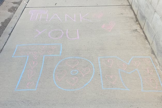 'Thank you Tom' written in pink and blue chalk on a footpath. 