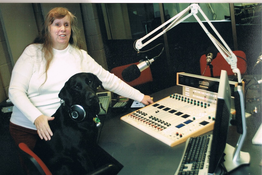 Elaine is standing with one arm on a radio control panel, one arm on her black guide dog.