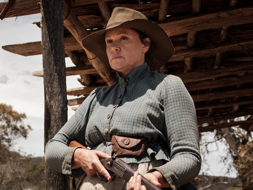 A woman in her 50s dressed in late 19th century skirt and shirt and stockman's hat, holding rifle, with porch of hut behind.