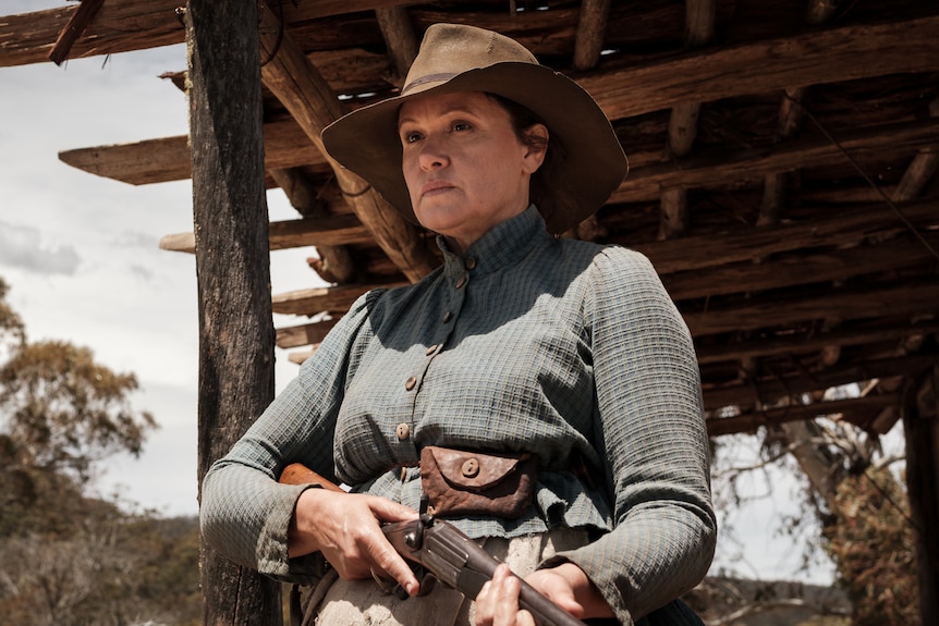A woman in her 50s dressed in late 19th century skirt and shirt and stockman's hat, holding rifle, with porch of hut behind.