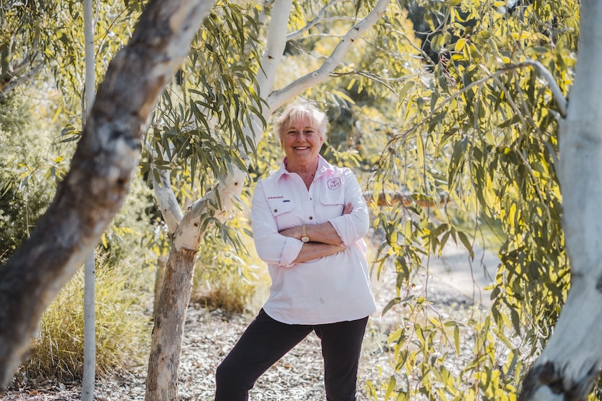 A woman stands with her arms crossed under a tree.